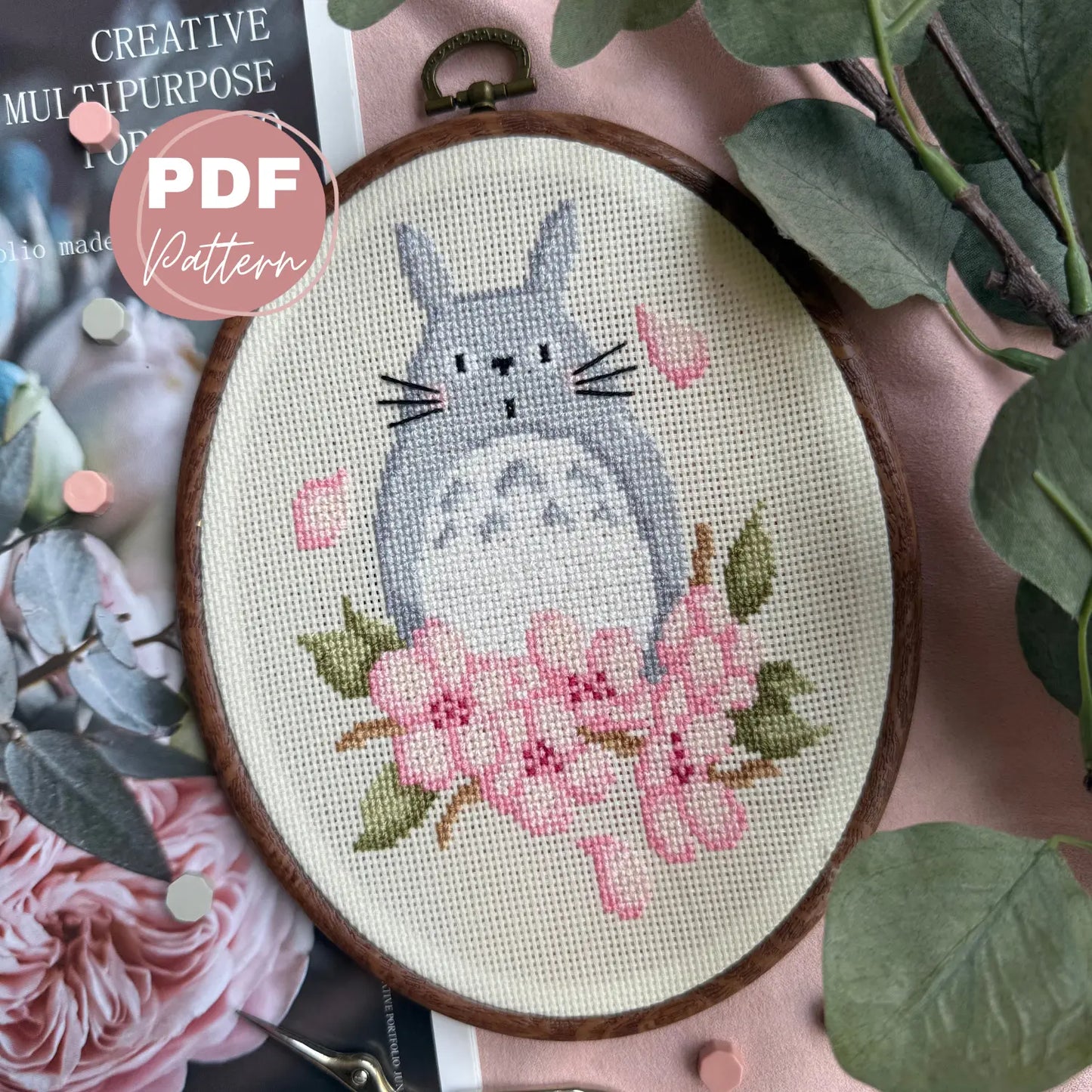 Cross stitched design of a cat totoro like creature with flowers sakura in an embroidery hoop, surrounded by green leaves and pink flowers.