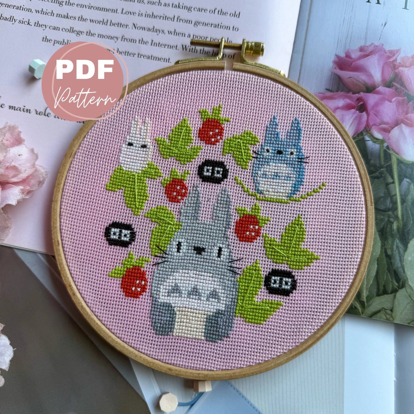 Cross stitched totoro studio ghibli design of cats with flowers in an embroidery hoop on a pink background.