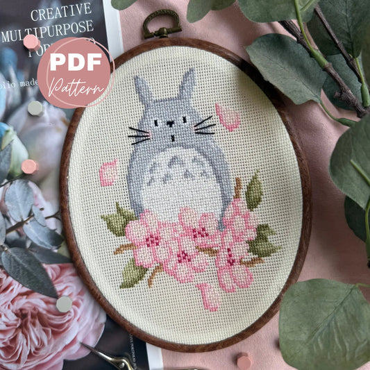 Cross stitched design of a cat totoro like creature with flowers sakura in an embroidery hoop, surrounded by green leaves and pink flowers.