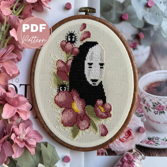 Cross stitched design of a character surrounded No face studio ghibli by flowers in an embroidery hoop, with a cup of tea and pink flowers in the background.