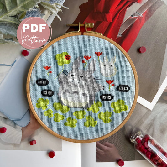 Embroidery hoop with cross stitched totoro studio ghibli  design of cartoon characters and flowers, surrounded by embroidery supplies.